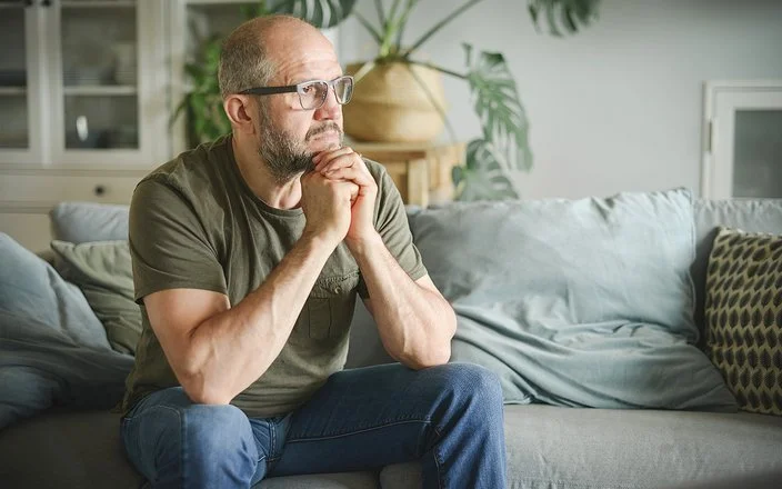 Ein Mann sitzt auf einer Couch und schaut nachdenklich zur Siete.