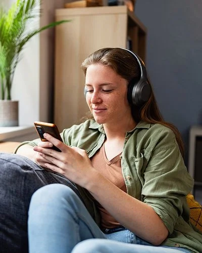 Eine junge Frau sitzt mit einem entspannten Lächeln auf einem Sofa, sie trägt große kabellose Kopfhörer und blickt auf ihr Smartphone.