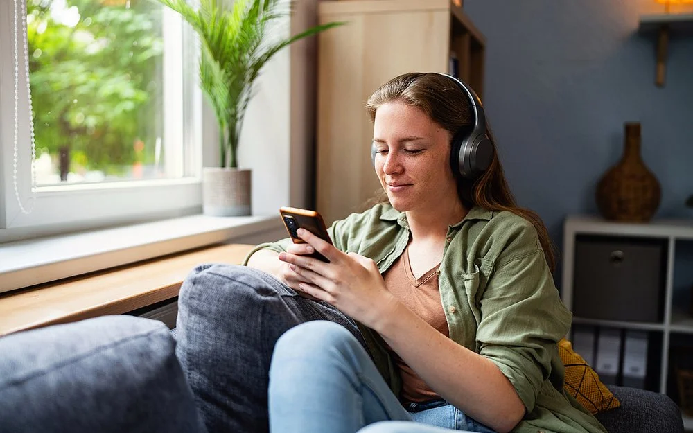 Eine junge Frau sitzt mit einem entspannten Lächeln auf einem Sofa, sie trägt große kabellose Kopfhörer und blickt auf ihr Smartphone.