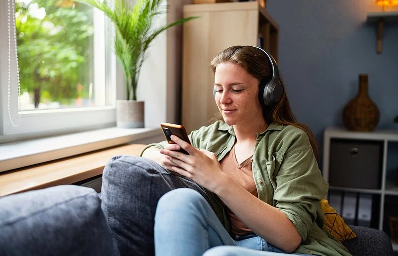 Eine junge Frau sitzt mit einem entspannten Lächeln auf einem Sofa, sie trägt große kabellose Kopfhörer und blickt auf ihr Smartphone.