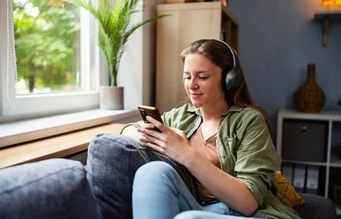 Eine junge Frau sitzt mit einem entspannten Lächeln auf einem Sofa, sie trägt große kabellose Kopfhörer und blickt auf ihr Smartphone.