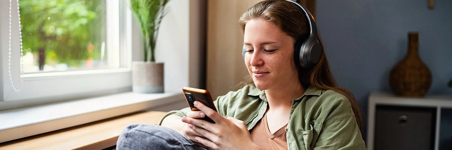 Eine junge Frau sitzt mit einem entspannten Lächeln auf einem Sofa, sie trägt große kabellose Kopfhörer und blickt auf ihr Smartphone.
