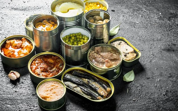Auf einer Schieferplatte stehen unterschiedliche geöffnete Konservendosen, unter anderem mit Ananas, Pilzen, Gemüse und Fisch.