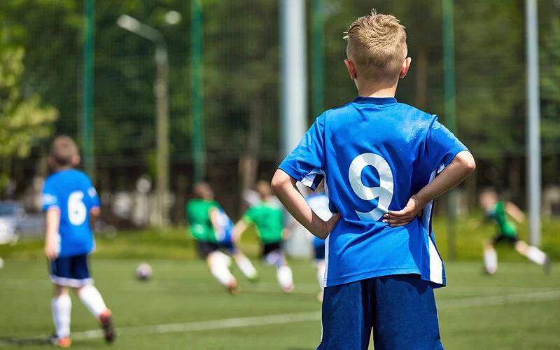 Im Vordergrund ist ein Junge im blauen Fußballtrikot von hinten zu sehen, der das Spielgeschehen seiner Mannschaft mit in die Hüfte gestemmtem Armen beobachtet.