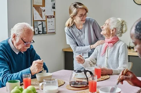 Eine Pflegerin begleitet ältere Menschen beim Frühstück. Sie steht am Tisch hinter einer älteren Frau und unterhält sich mit dieser. Mit am Tisch sitzen noch zwei ältere Männer, die ihr Frühstück aus Schüsseln essen. Auf dem Tisch steht eine Kaffeekanne, eine Milchkanne, Saft und Obst.