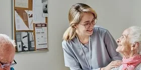 Eine Pflegerin begleitet ältere Menschen beim Frühstück. Sie steht am Tisch hinter einer älteren Frau und unterhält sich mit dieser. Mit am Tisch sitzen noch zwei ältere Männer, die ihr Frühstück aus Schüsseln essen. Auf dem Tisch steht eine Kaffeekanne, eine Milchkanne, Saft und Obst.