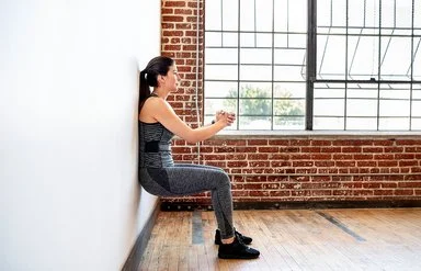 Eine Frau praktiziert Wandpilates. Sie steht mit dem Rücken an der Wand, beugt die Knie und presst ihre Hände vor der Brust zusammen.