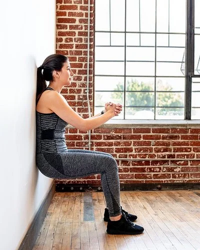 Eine Frau praktiziert Wandpilates. Sie steht mit dem Rücken an der Wand, beugt die Knie und presst ihre Hände vor der Brust zusammen.