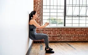 Eine Frau praktiziert Wandpilates. Sie steht mit dem Rücken an der Wand, beugt die Knie und presst ihre Hände vor der Brust zusammen.