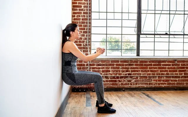 Eine Frau praktiziert Wandpilates. Sie steht mit dem Rücken an der Wand, beugt die Knie und presst ihre Hände vor der Brust zusammen.