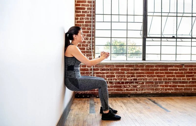 Eine Frau praktiziert Wandpilates. Sie steht mit dem Rücken an der Wand, beugt die Knie und presst ihre Hände vor der Brust zusammen.