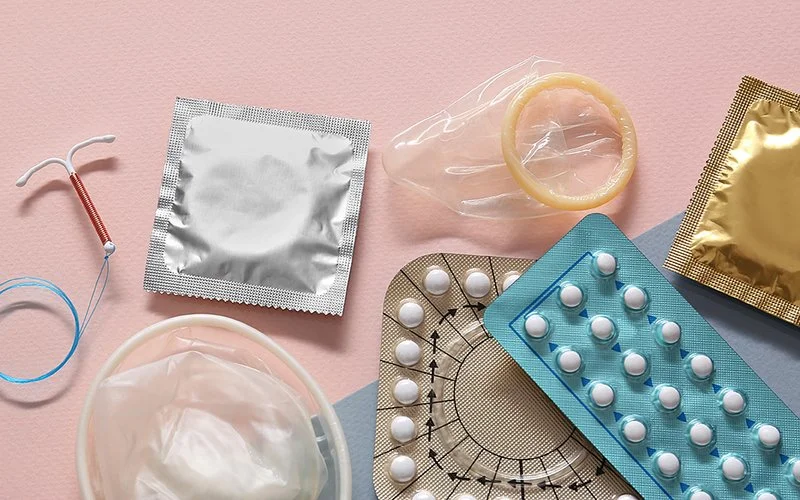 Verschiedene Verhütungsmittel liegen auf rosa Hintergrund, darunter Kondome, ein Femidom, Antibabypillen, eine Spirale und ein Verhütungsring.