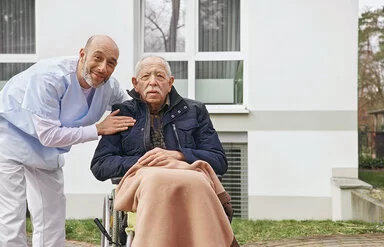 Ein älterer Mann mit dunkelblauer Jacke sitzt in einem Rollstuhl auf einer Terrasse, auf seinen Beinen liegt eine Decke. Eine Pflegefachkraft in hellblauer Kleidung beugt sich zum Mann herunter, eine Hand ruht auf der Schulter des Mannes. Beide lächeln in die Kamera.