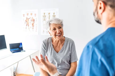 Eine ältere Patientin sitzt lachend vor ihrem Arzt und lässt sich beraten.