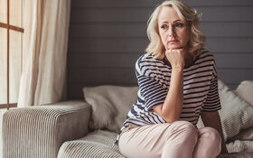Eine Frau, die unter chronischen Schmerzen leidet, sitzt mit traurigem Blick auf dem Sofa.