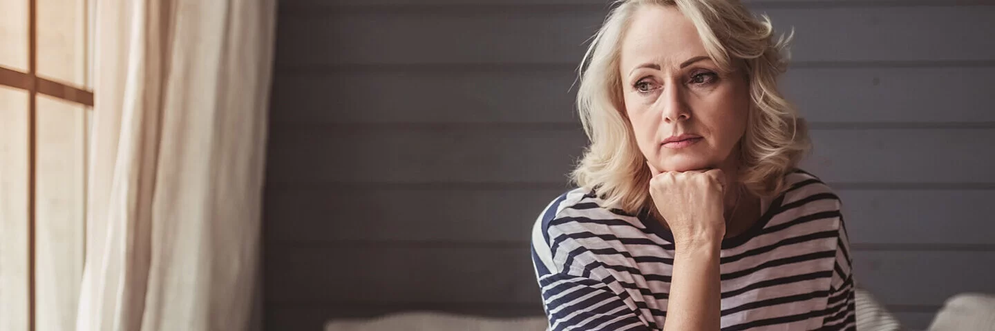 Eine Frau, die unter chronischen Schmerzen leidet, sitzt mit traurigem Blick auf dem Sofa.