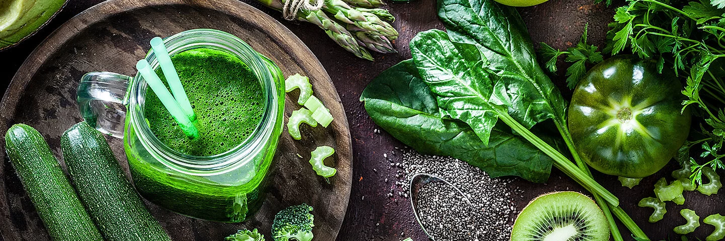 Ein frischer grüner Smoothie im Glas, umgeben von frischem Gemüse wie Zucchini, Spinat und Paprika.