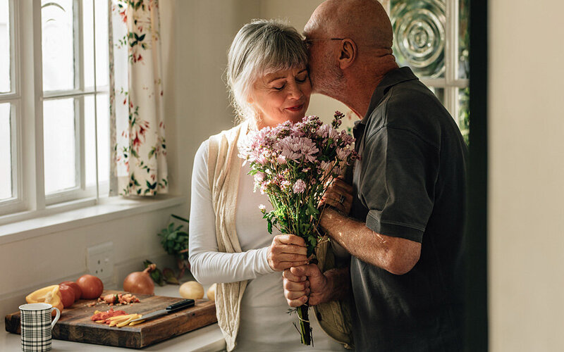 Ein Mann schenkt seiner Frau einen Blumenstrauß und küsst sie sanft auf die Wange.