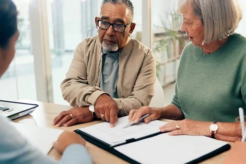 Ein älteres Paar befindet sich in einem Büro einer Beratungsstelle. Sie sitzen an einem Schreibtisch und haben vor sich mehrere Dokumente liegen, die sie gemeinsam mit einer Beraterin besprechen, die ihnen gegenüber sitzt.