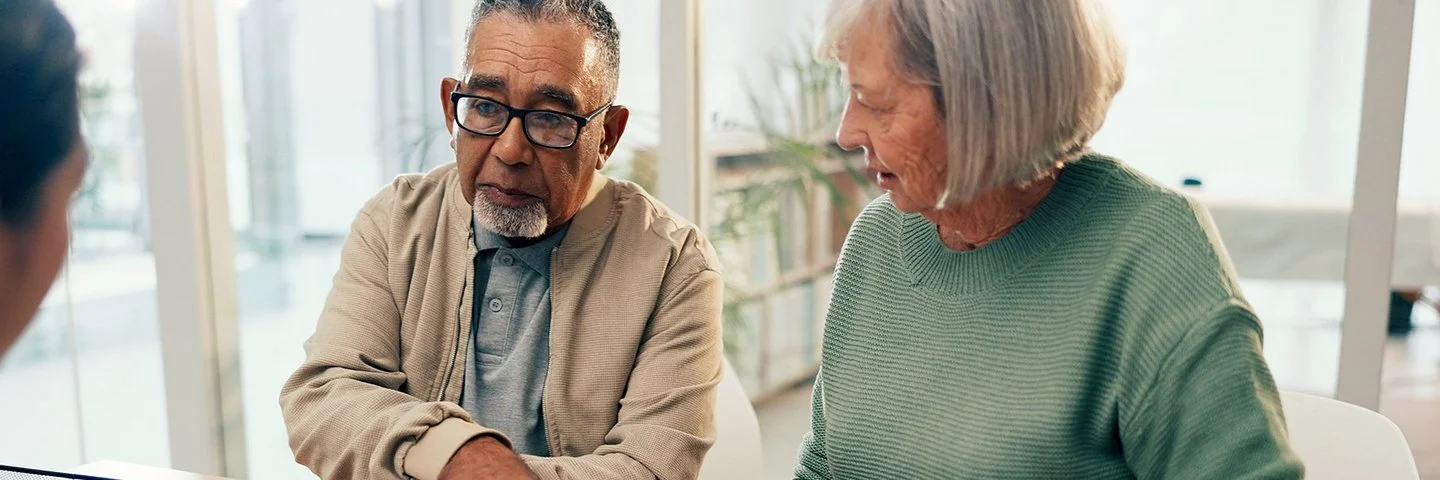 Ein älteres Paar befindet sich in einem Büro einer Beratungsstelle. Sie sitzen an einem Schreibtisch und haben vor sich mehrere Dokumente liegen, die sie gemeinsam mit einer Beraterin besprechen, die ihnen gegenüber sitzt.