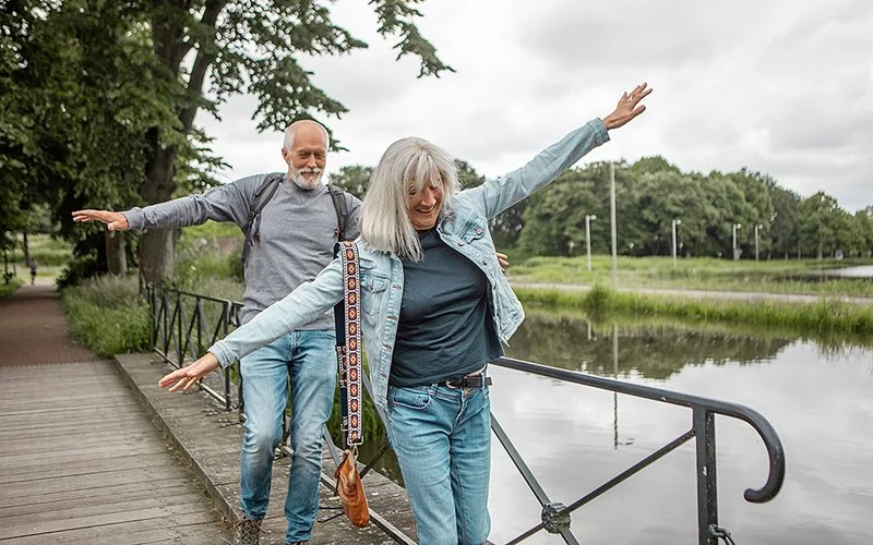 Eine Mann und eine Frau mit grauen Haaren balancieren lachend auf der Bordsteinkante einer Brücke.