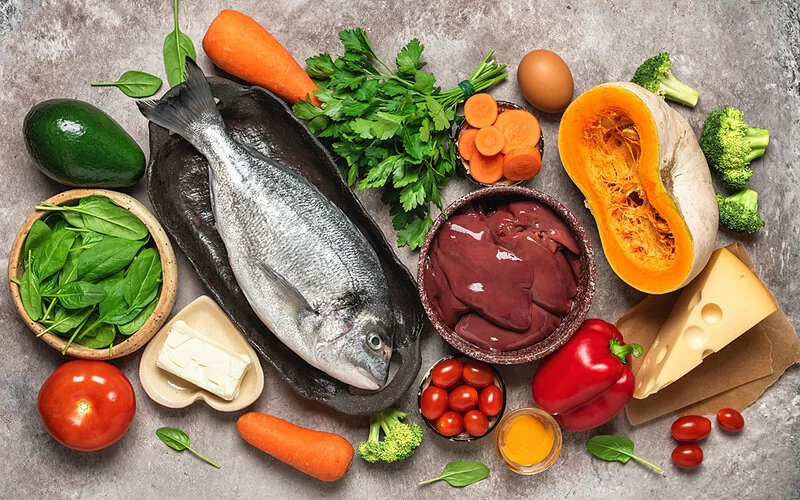 Verschiedene frische Lebensmittel, die Vitamin A oder die Vorstufe Betacarotin enthalten, darunter Leber, Ei, Käse, Karotten und Spinat.