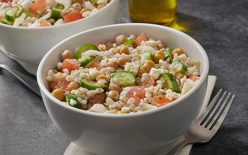 Zwei Schüsseln mit einem Salat aus Graupen, Gurken, Tomaten und Kichererbsen stehen auf einem Tisch.