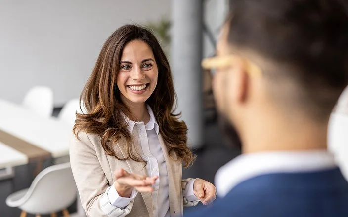 Eine lächelnde Frau im Business-Outfit spricht mit einem Mann, der unscharf im Vordergrund zu sehen ist.