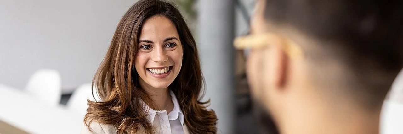 Eine lächelnde Frau im Business-Outfit spricht mit einem Mann, der unscharf im Vordergrund zu sehen ist.