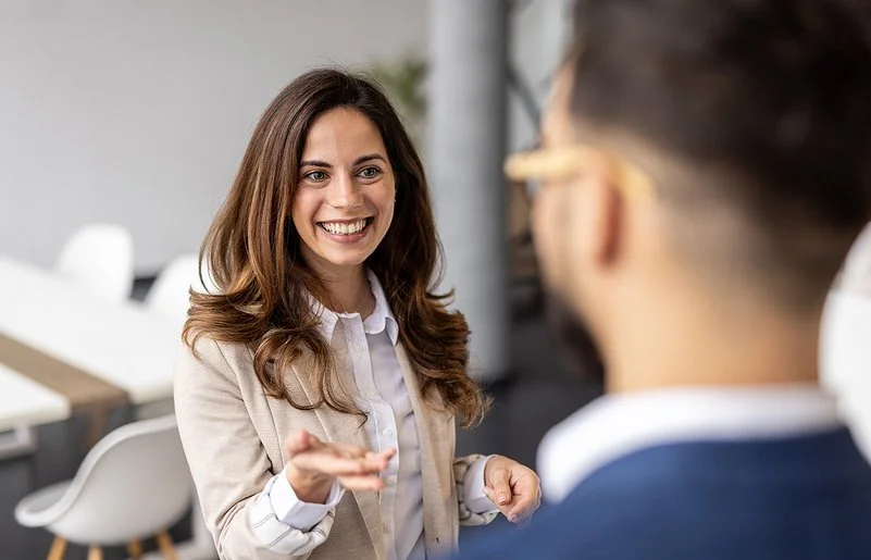 Eine lächelnde Frau im Business-Outfit spricht mit einem Mann, der unscharf im Vordergrund zu sehen ist.