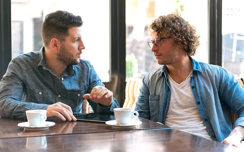 Zwei Männer sitzen an einem Tisch, haben jeweils eine Tasse Kaffee vor sich stehen und unterhalten sich angeregt. Zwischen ihnen liegt ein Tablet.