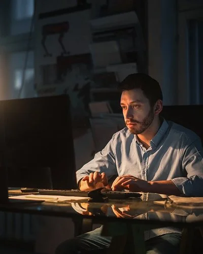 In einem dunklen Büro sitzt ein Mann an einem Schreibtisch, der durch eine Tischleuchte beleuchtet wird. Er tippt auf einer Tastatur und schaut auf den Monitor.