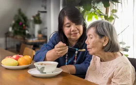 Eine pflegende Angehörige hilft ihrer alten Mutter beim Abendessen.