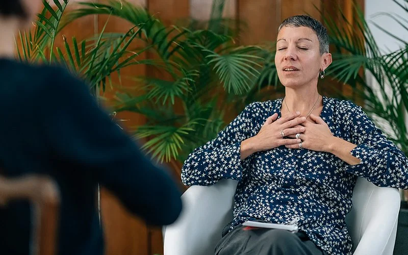 Eine Frau sitzt auf einem Stuhl einer anderen Frau gegenüber. Von letzterer ist lediglich ein unscharfer Ausschnitt der Rückansicht zu sehen. Die Frau hat die Augen geschlossen und beide Hände auf den oberen Brustkorb gelegt. Ihr Mund ist geöffnet.