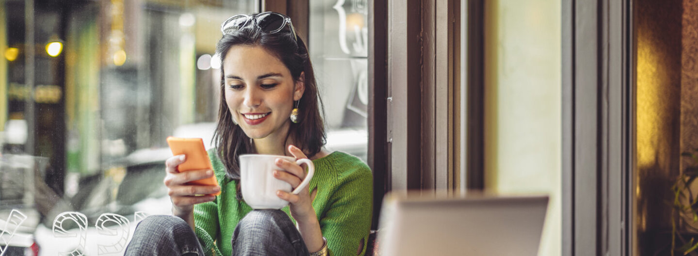 Eine junge Frau sitzt in einem Café neben ihrem geöffneten Laptop und trinkt ein Heißgetränk während sie auf ihr Smartphone schaut.