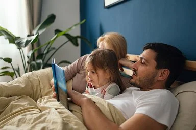 Eine junge Familie mit einem kleinen Kind liegt gemeinsam in einem gemütlichen Bett im Elternschlafzimmer. Der Vater liest seiner Tochter aus einem Buch vor. Das kleine Mädchen hört gespannt zu.