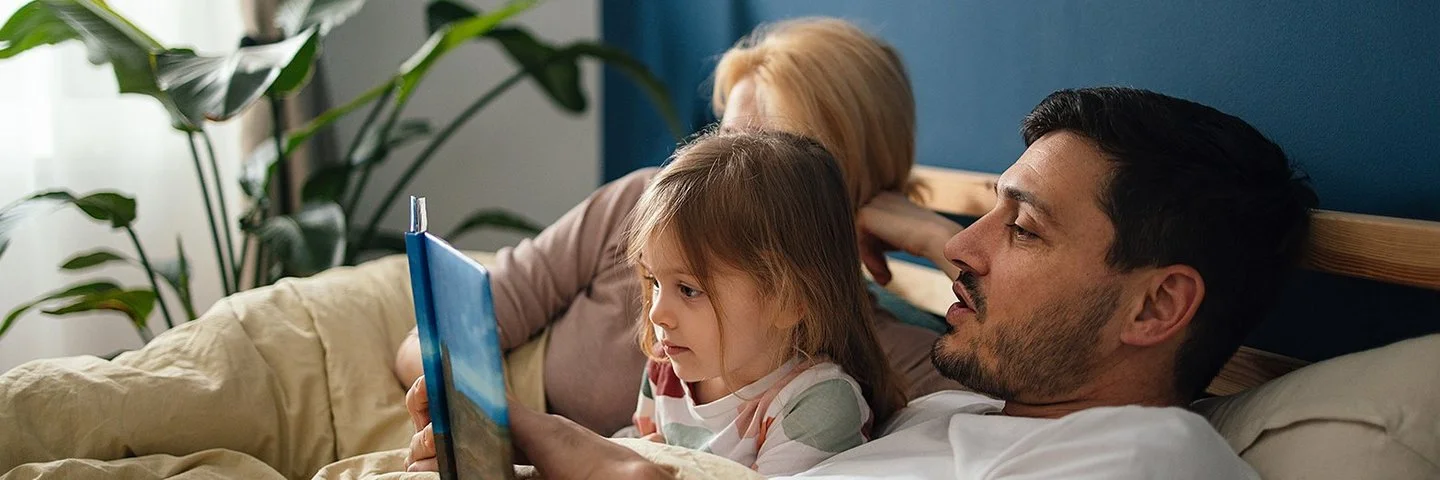 Eine junge Familie mit einem kleinen Kind liegt gemeinsam in einem gemütlichen Bett im Elternschlafzimmer. Der Vater liest seiner Tochter aus einem Buch vor. Das kleine Mädchen hört gespannt zu.