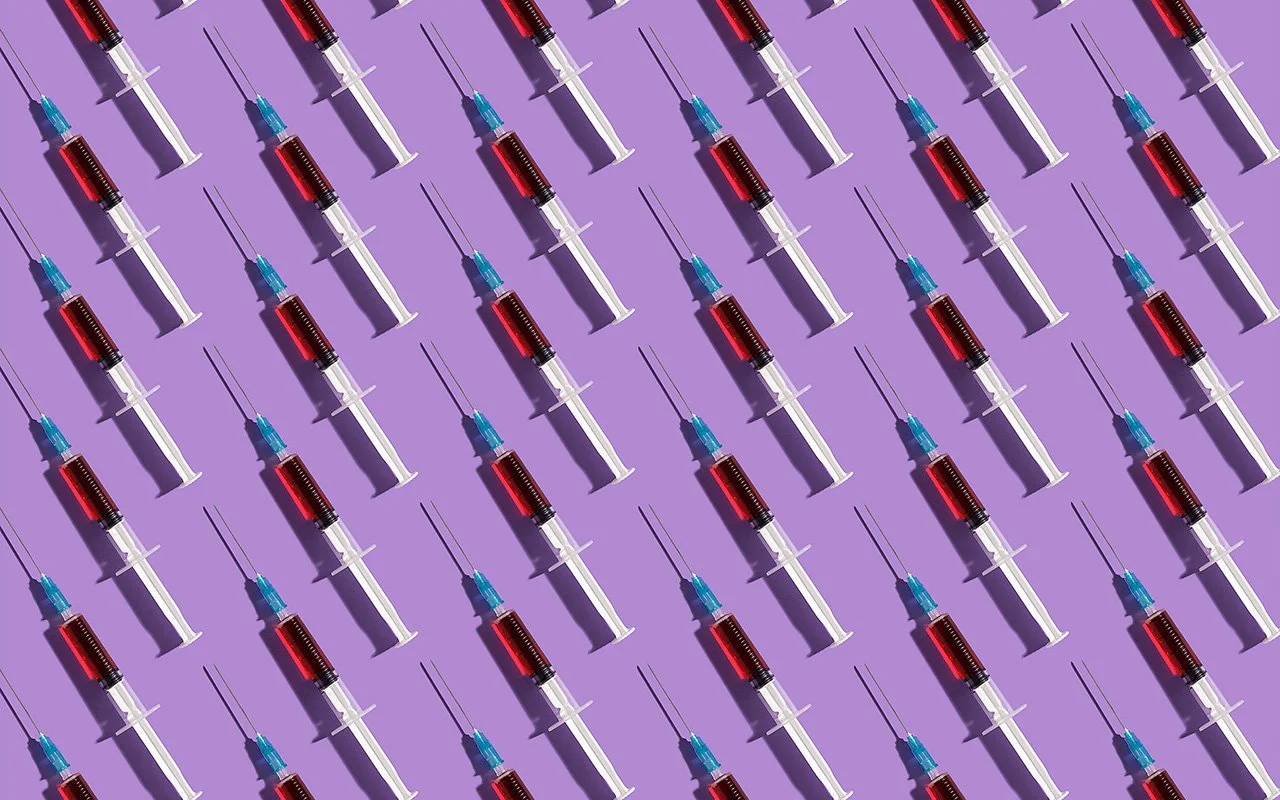 Mehrere Spritzen mit rotem Inhalt und blauen Kappen liegen diagonal auf einer lilafarbenen Fläche.