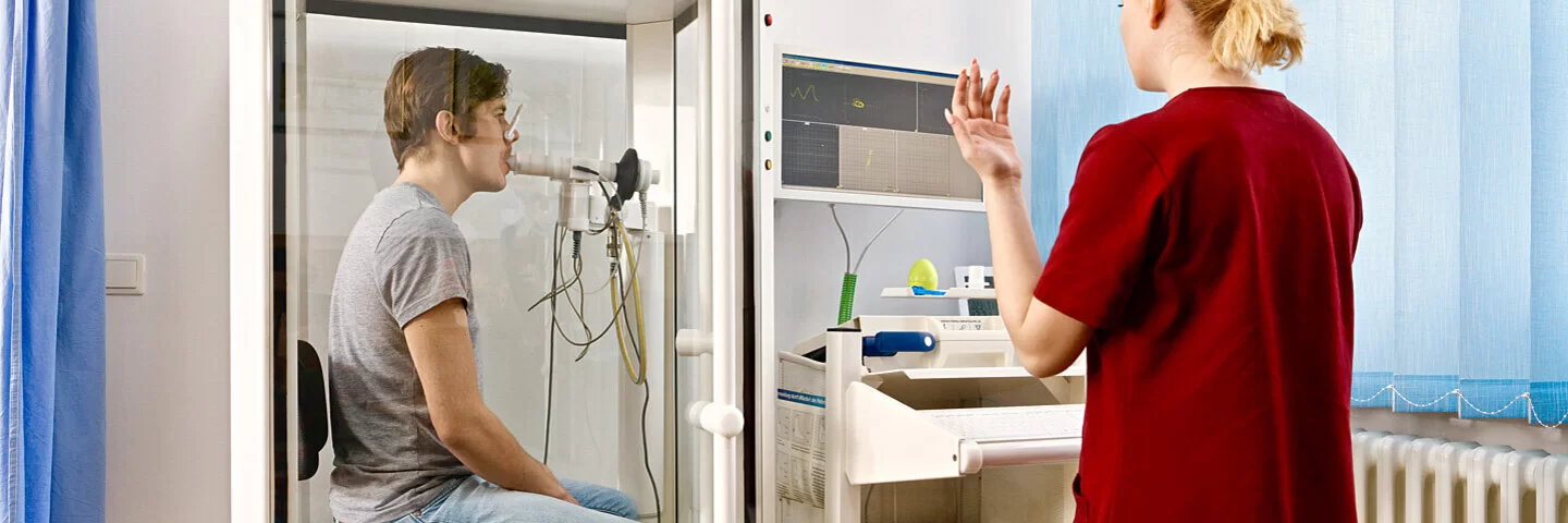 Ein Mann sitzt in einer Kabine, eine Pneumologin überwacht am Bildschirm die Lungenfunktion.