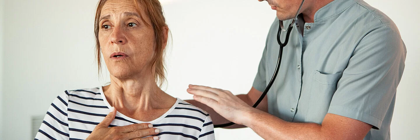 Eine ältere Frau mit COPD und Atemnot ist beim Arzt, der ihre Lunge abhorcht.