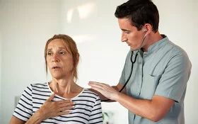 Eine ältere Frau mit COPD und Atemnot ist beim Arzt, der ihre Lunge abhorcht.