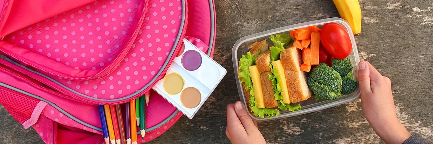 Eine rosafarbene Schultasche mit weißen Punkten, die auf einem Holztisch liegt. Aus der Schultasche ragen Buntstifte und ein Tuschkasten hervor. Daneben liegen ein gelber und ein roter Apfel sowie eine Banane. Eine Person hält eine transparente Brotdose ins Bild, die mit gesunden Pausenbroten und frischem Gemüse gefüllt ist.