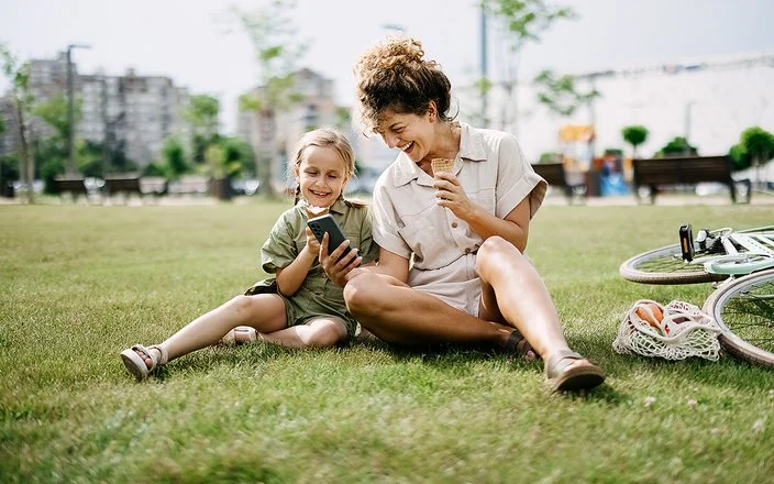 Eine Frau hält einem Mädchen lachend ein Smartphone hin, beide sitzen im Gras.