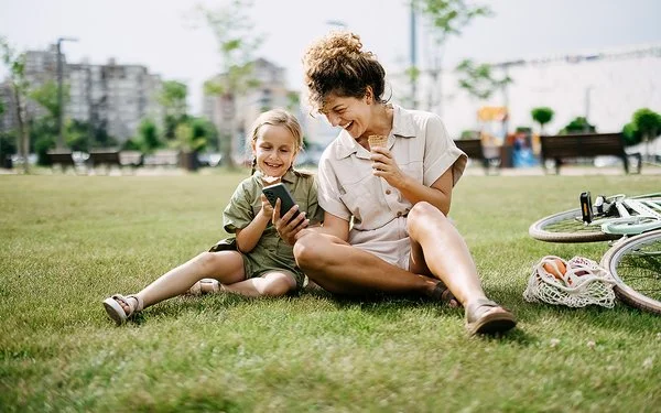 Eine Frau hält einem Mädchen lachend ein Smartphone hin, beide sitzen im Gras.