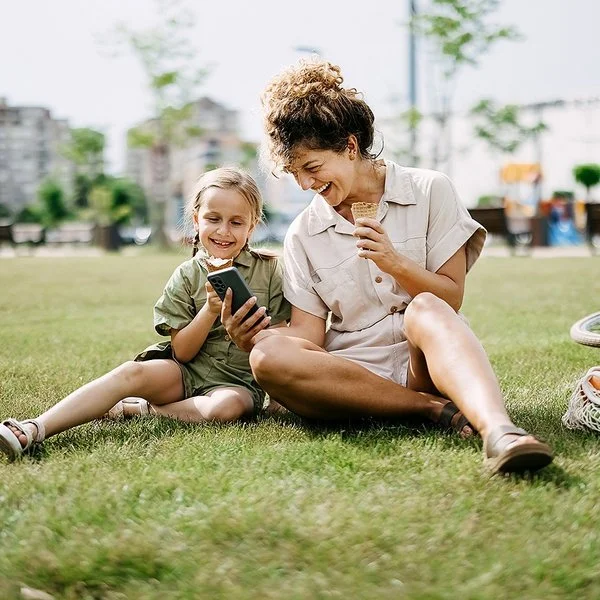 Eine Frau hält einem Mädchen lachend ein Smartphone hin, beide sitzen im Gras.
