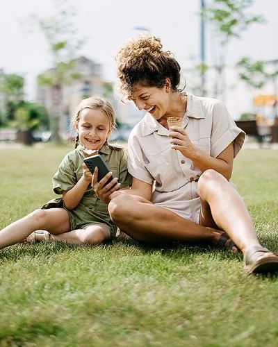 Eine Frau hält einem Mädchen lachend ein Smartphone hin, beide sitzen im Gras.