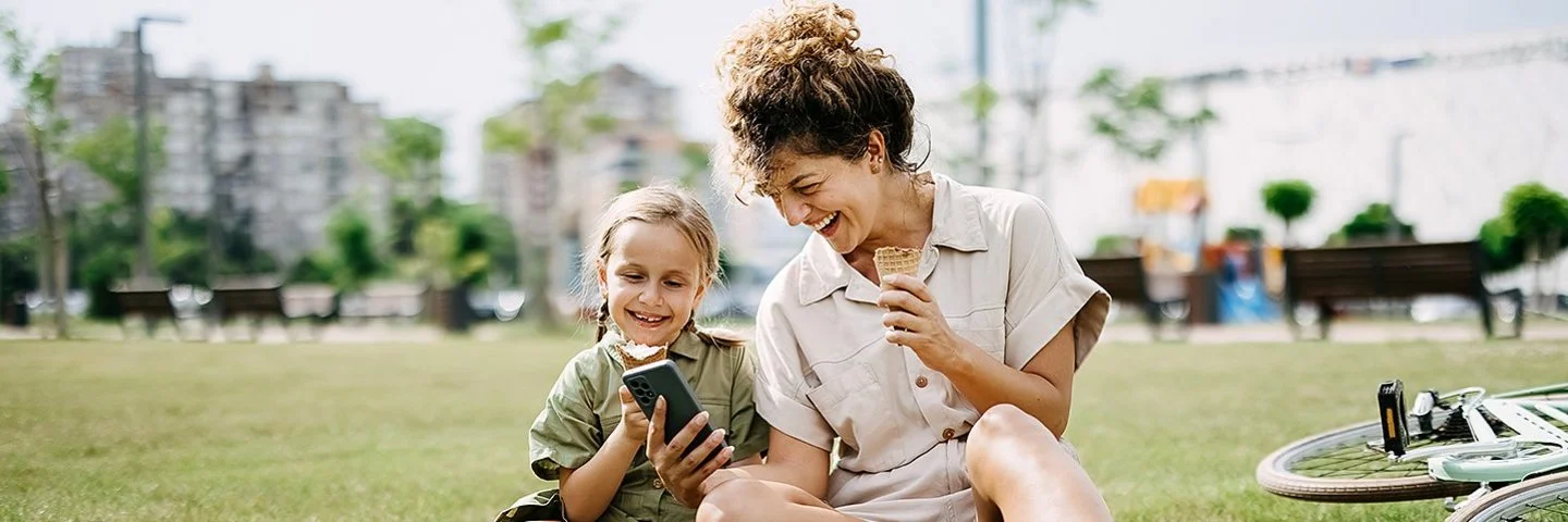 Eine Frau hält einem Mädchen lachend ein Smartphone hin, beide sitzen im Gras.