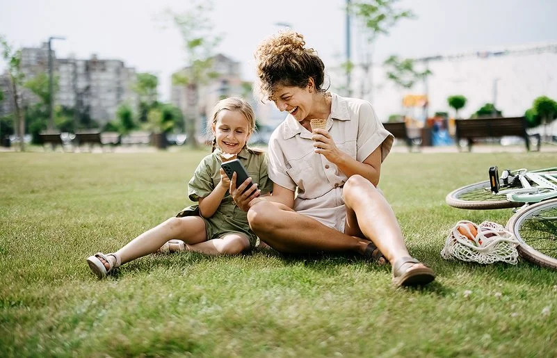 Eine Frau hält einem Mädchen lachend ein Smartphone hin, beide sitzen im Gras.