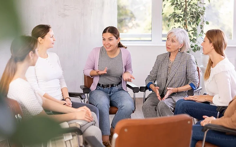 Eine Gruppe von Frauen unterschiedlichen Alters sitzt in einem Stuhlkreis. Eine Teilnehmerin spricht, während die anderen aufmerksam zuhören.