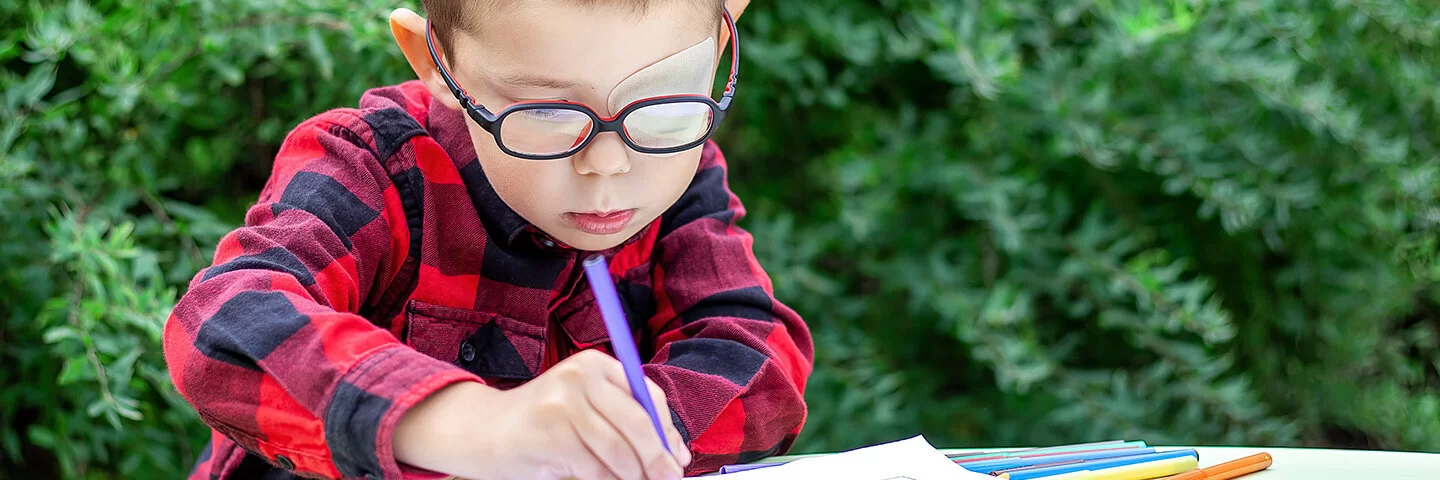 Ein Junge mit Amblyopie hat sein linkes Auge mit einem Okkluder abgeklept, er trägt dazu eine Brille und sitzt malend im Garten an einem Tisch.